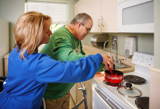 A caregiver assists an elderly person with cooking in the kitchen.