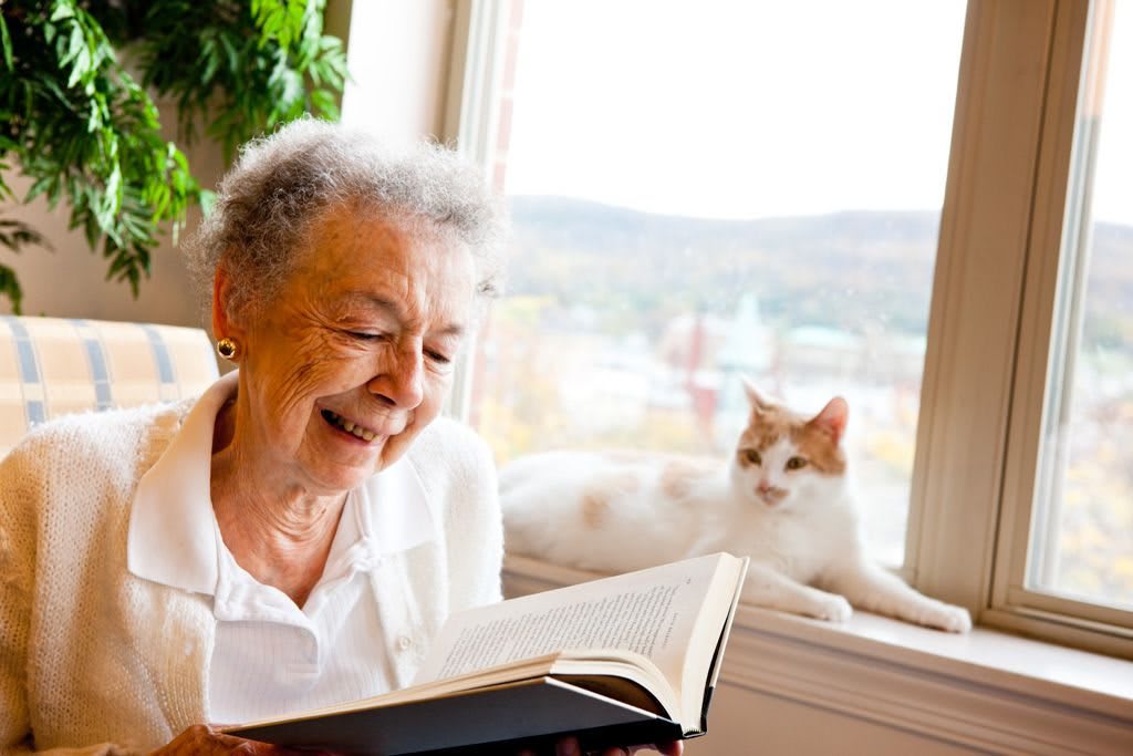 An elderly woman smiling while reading a book near a window with a cat lounging beside her.