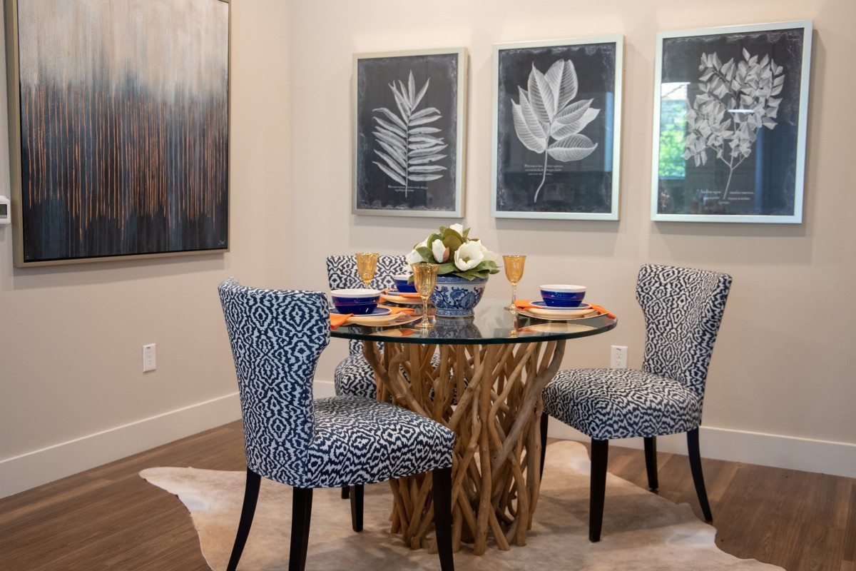 A modern dining room with a glass-top table and patterned chairs, complemented by botanical artwork on the walls.