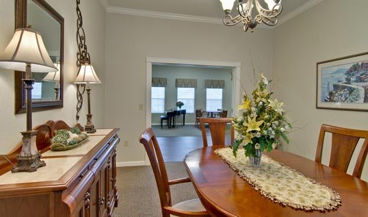 A dining room with a wooden table and chairs, featuring a floral centerpiece and a chandelier. In the background, there's a view of the living room with large windows and a piano.