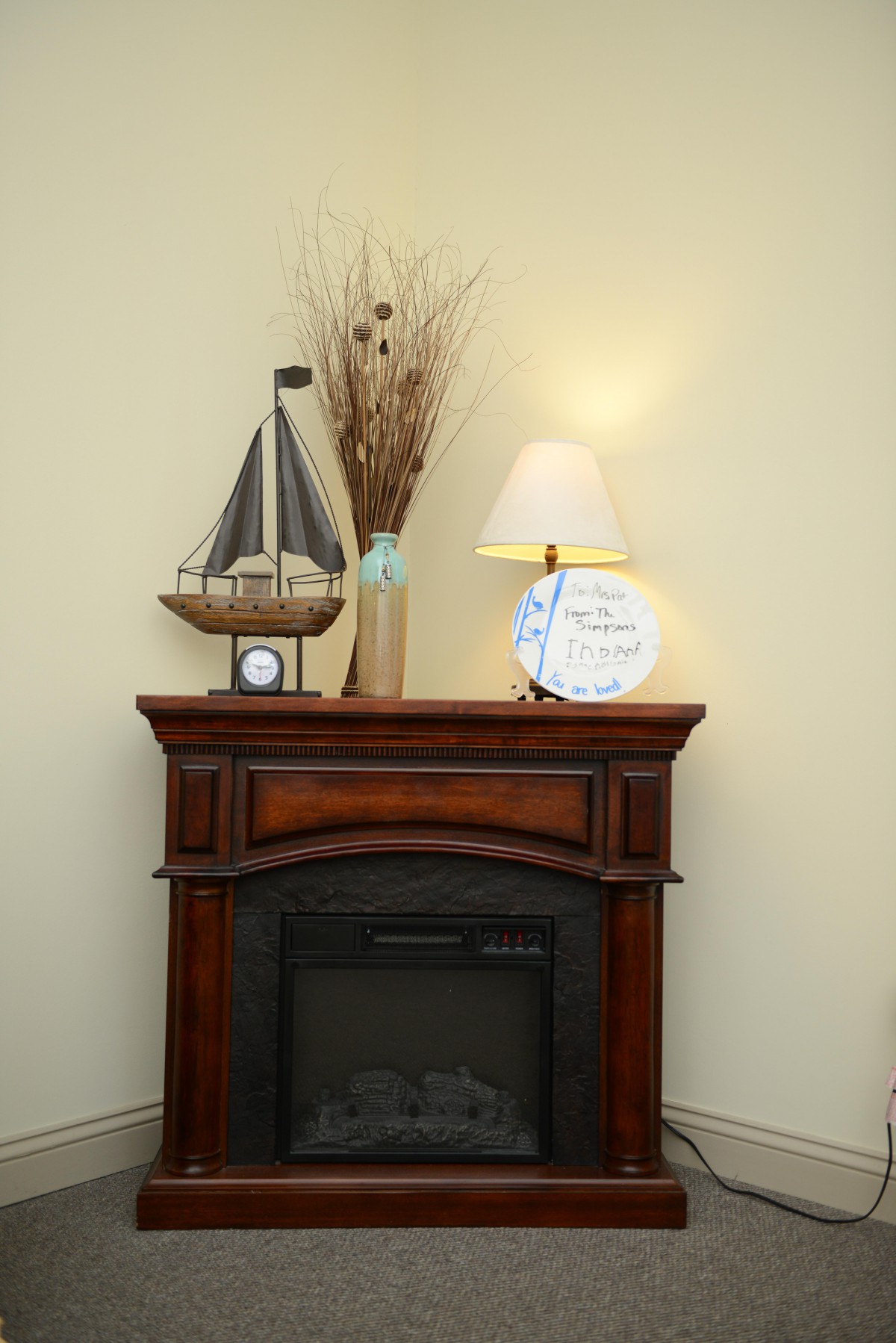 A cozy fireplace mantle decorated with a model sailboat, a lamp, a decorative plate, and a vase with dried branches.