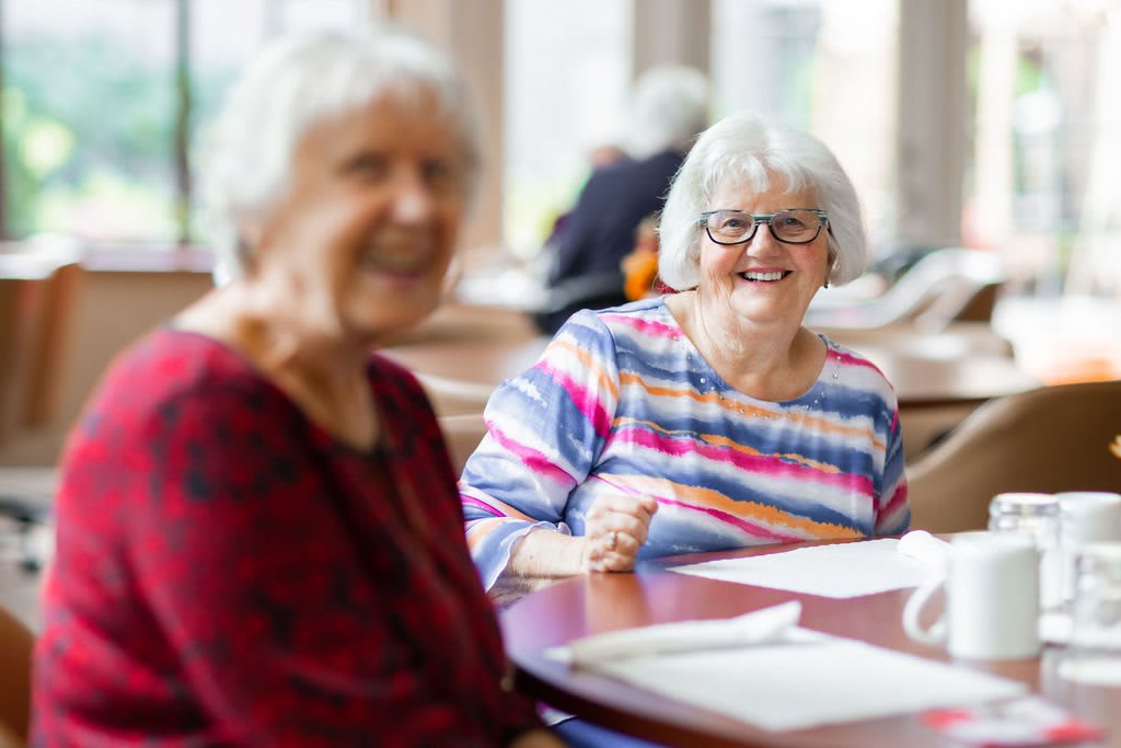 Two elderly women smiling and enjoying a conversation indoors.