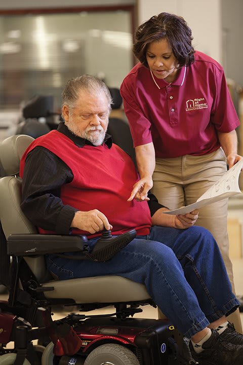 An elderly man in a wheelchair receives assistance from a caregiver while using a tablet.