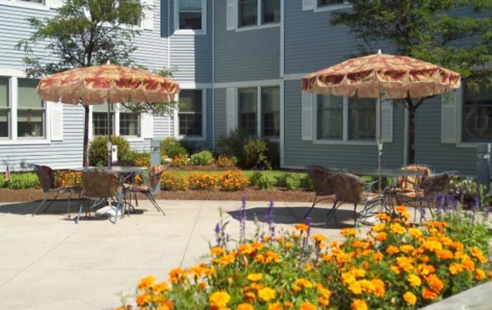 A sunny outdoor patio with colorful flowers and seating under umbrellas next to a residential building.