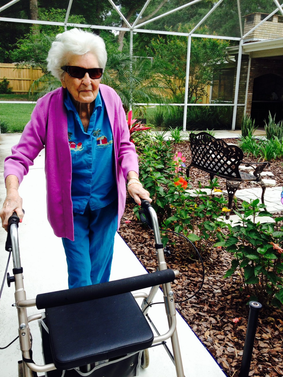 An elderly woman walking outside using a walker, enjoying the garden scenery.