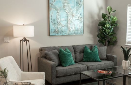 A cozy living room with a gray sofa adorned with teal cushions, a piece of abstract artwork on the wall, and a potted plant in the corner.