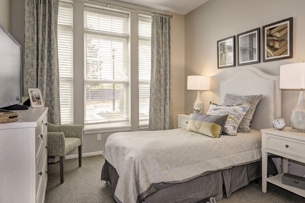 A cozy bedroom with a neatly made bed, large windows, and soft lighting.