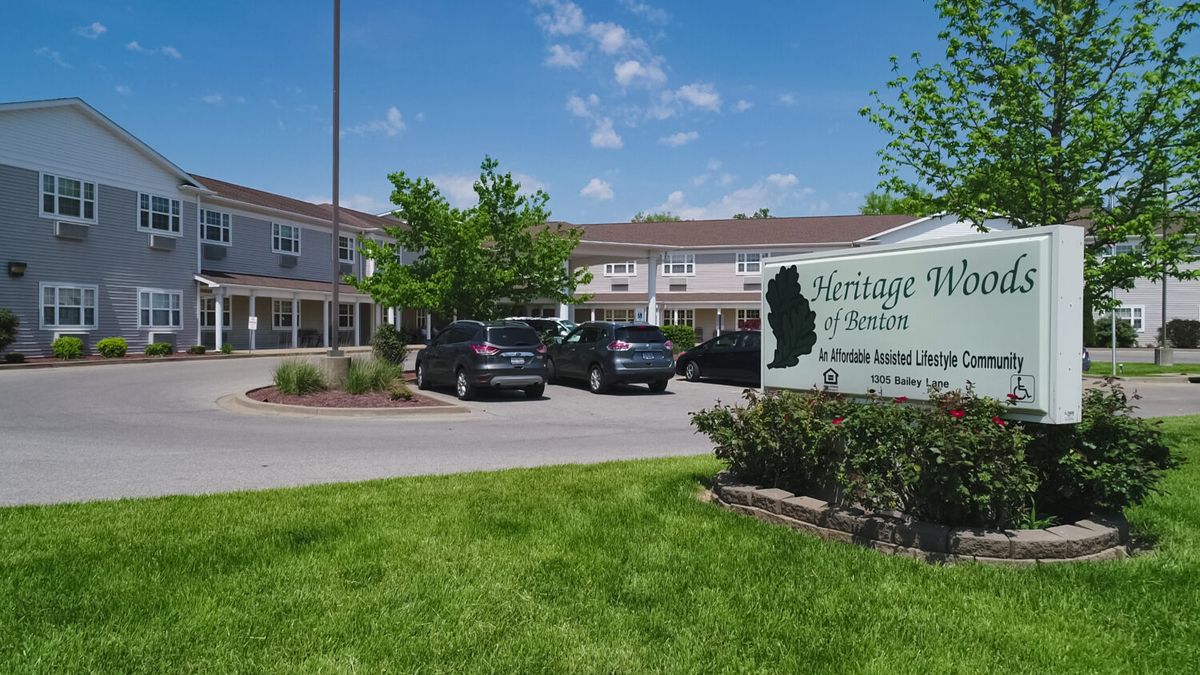 A residential building with a parking lot and sign, surrounded by greenery.