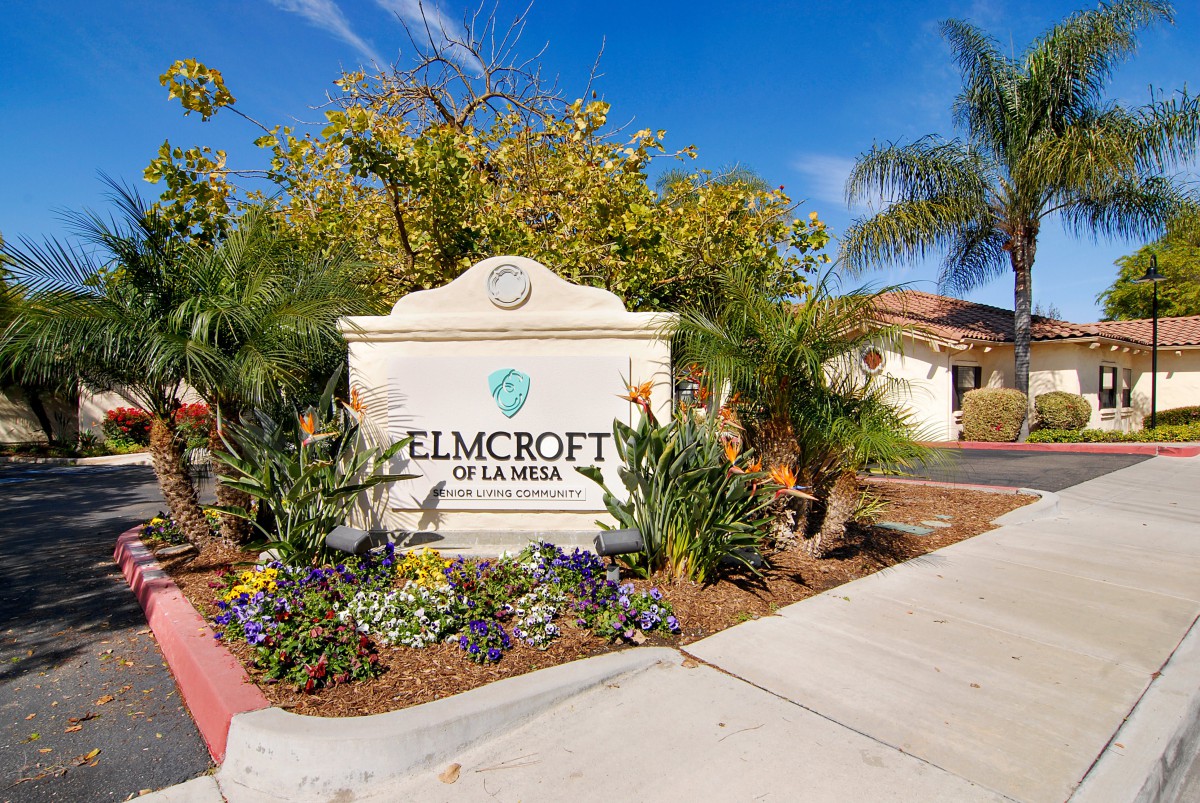 Entrance sign of Elmcroft of La Mesa surrounded by palm trees and flowers.