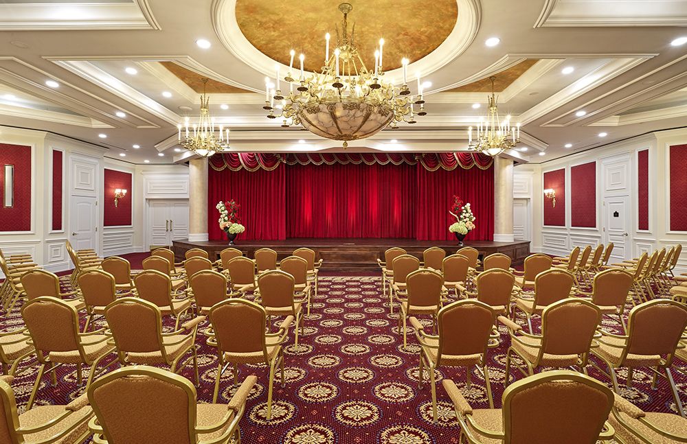 An elegant auditorium with red curtains and ornate chandeliers, featuring rows of comfortable seating.