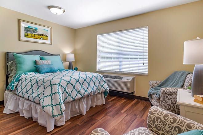 A cozy bedroom with a neatly made bed, featuring a patterned comforter, wooden flooring, and a window with blinds.