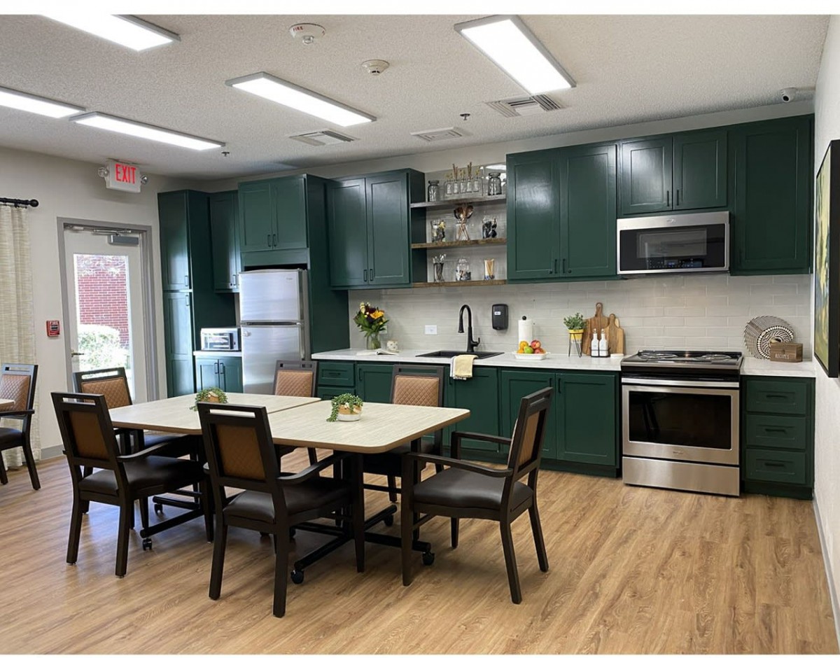 A modern kitchen and dining area featuring green cabinets, a wooden table with chairs, stainless steel appliances, and wood flooring.