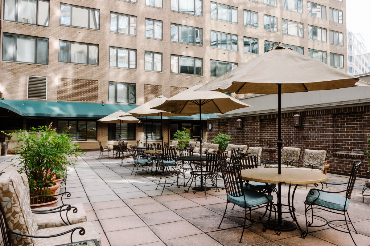 An outdoor patio area with tables, chairs, and umbrellas surrounded by a multi-story building.