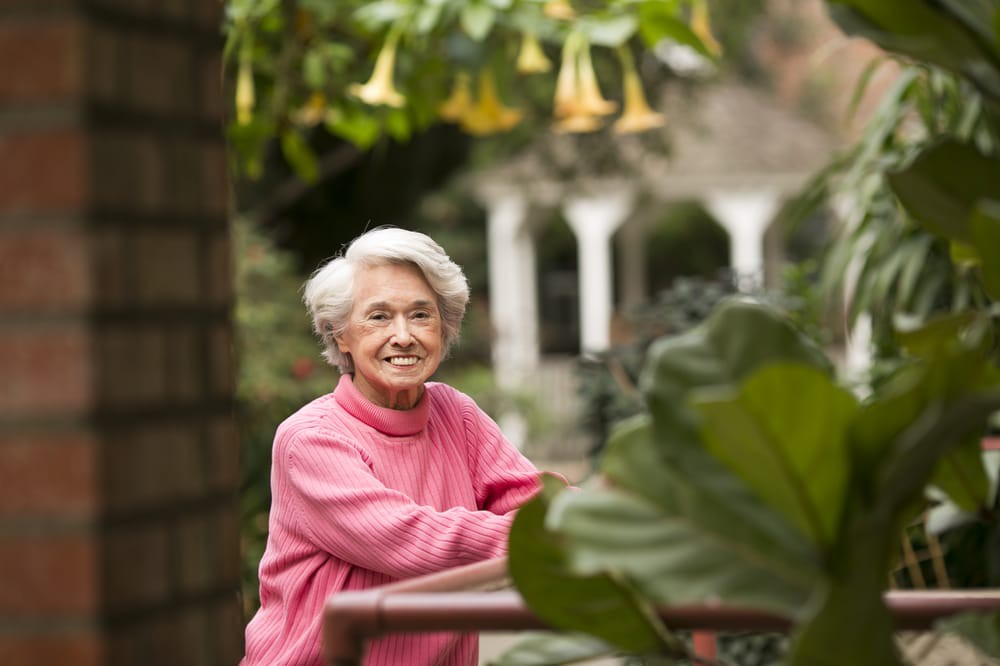 An elderly woman smiling in a garden surrounded by flowers.