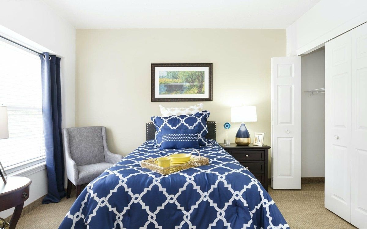 A cozy bedroom with a single bed featuring blue patterned bedding, a nightstand with a lamp, and an armchair.