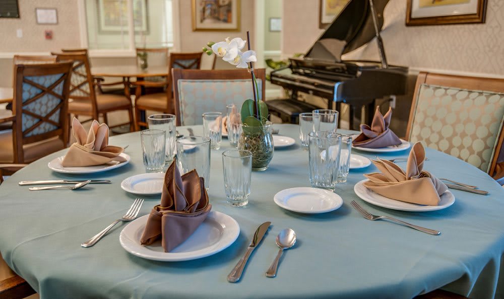 A dining table elegantly set with glasses, plates, silverware, and folded napkins in a room featuring a grand piano.