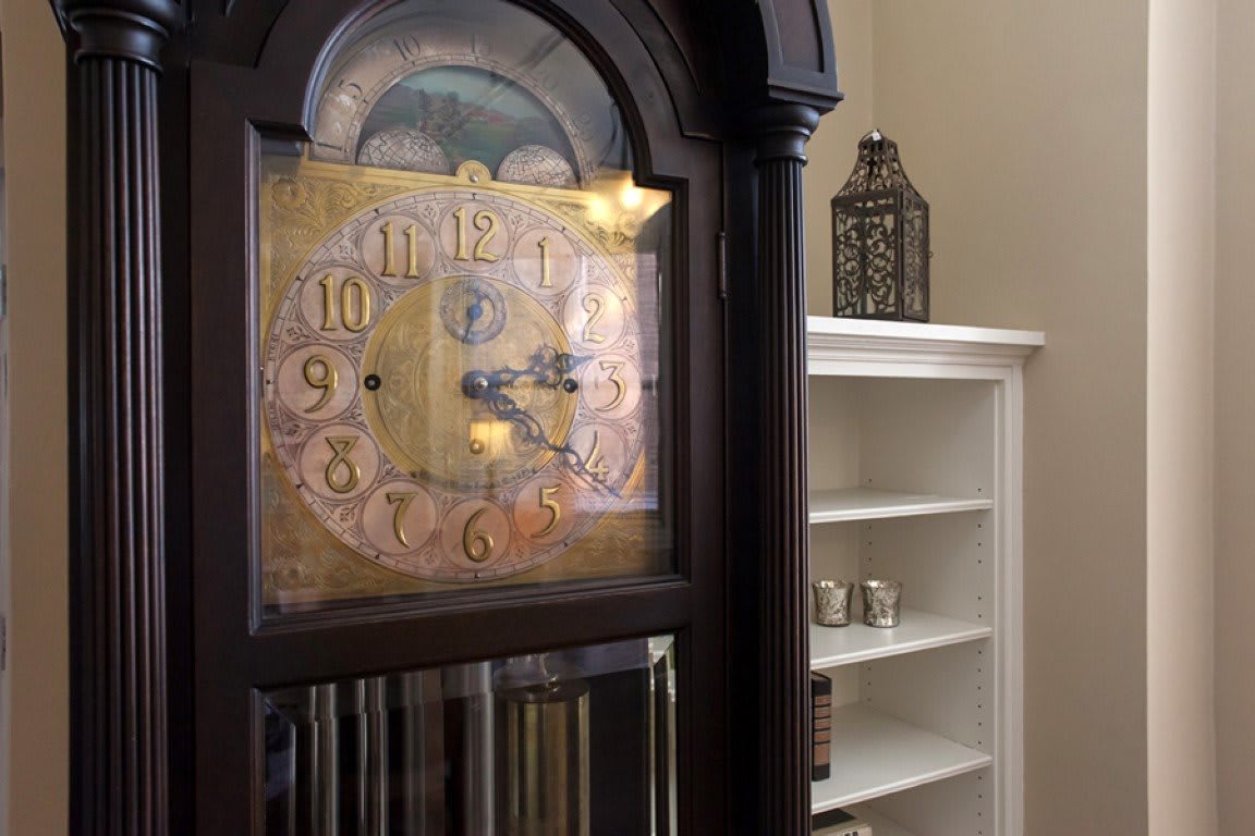 A vintage grandfather clock stands next to a white bookshelf with decorative items.