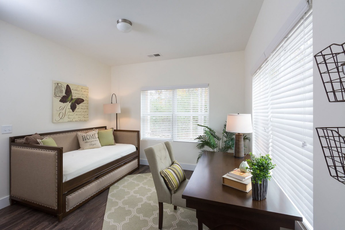 A cozy bedroom with a daybed, desk, and natural light from a large window.