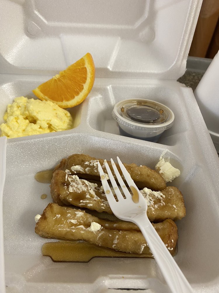 A takeout breakfast featuring pancakes with syrup, scrambled eggs, and an orange slice.