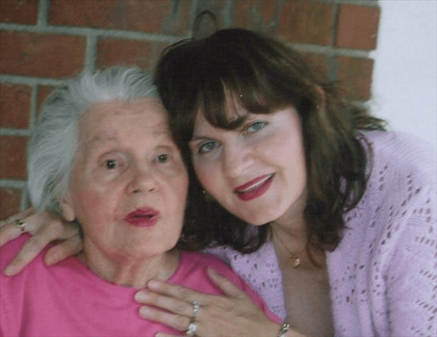 A middle-aged woman embraces an elderly woman in front of a brick wall.