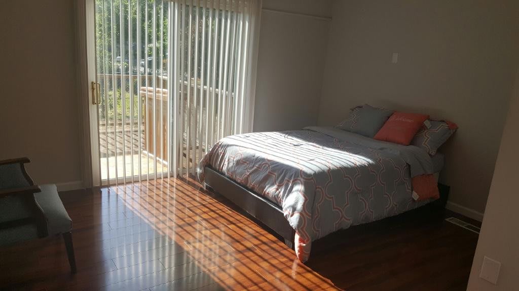 A sunlit bedroom with a bed and hardwood floors, featuring large windows with blinds.
