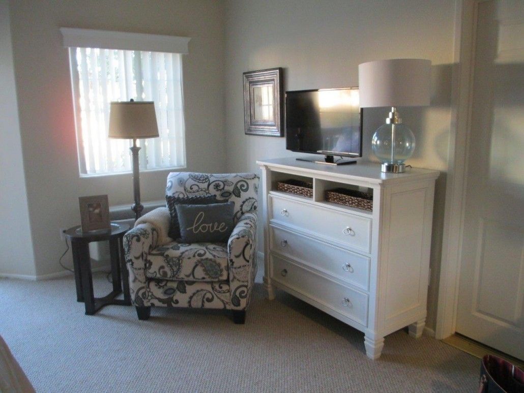 A cozy living room corner with a patterned armchair, side table with a lamp, dresser with a television on top, and a window letting in natural light.