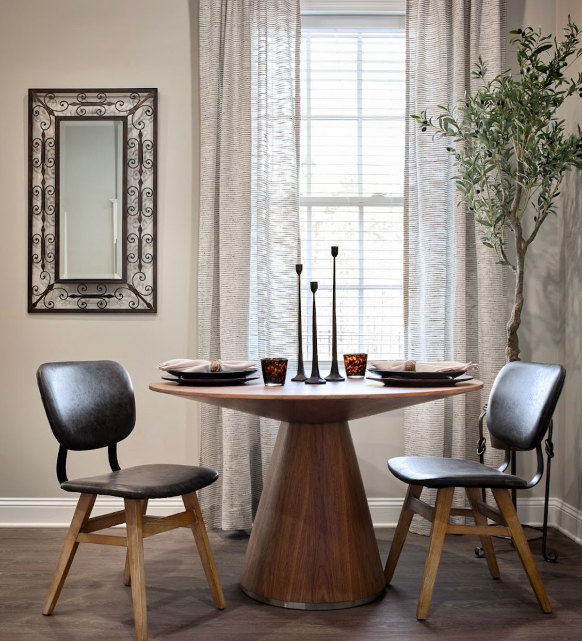 A cozy dining area featuring a round wooden table with two modern chairs, accented by a decorative mirror and tall candles.
