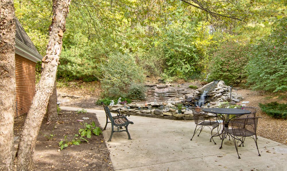 A serene patio with metal furniture overlooks a small waterfall surrounded by lush greenery.