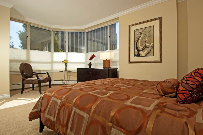 A modern bedroom with a large bed, geometric-patterned bedspread, and contemporary furniture, featuring an artwork on the wall and a corner window allowing natural light.