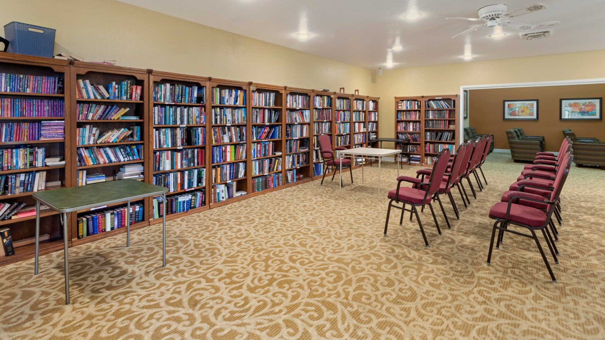A library room featuring bookshelves lined with numerous books, several chairs, and a few tables arranged in the center.