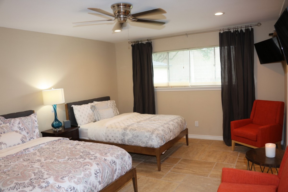 A well-lit bedroom featuring two beds, a lamp on the side, a ceiling fan, and dark curtains framing a window.