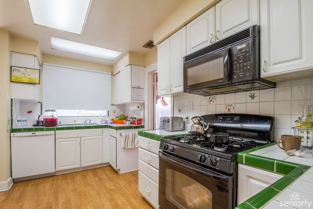 A cozy kitchen featuring a stove, microwave, and white cabinets with green-tiled countertops.