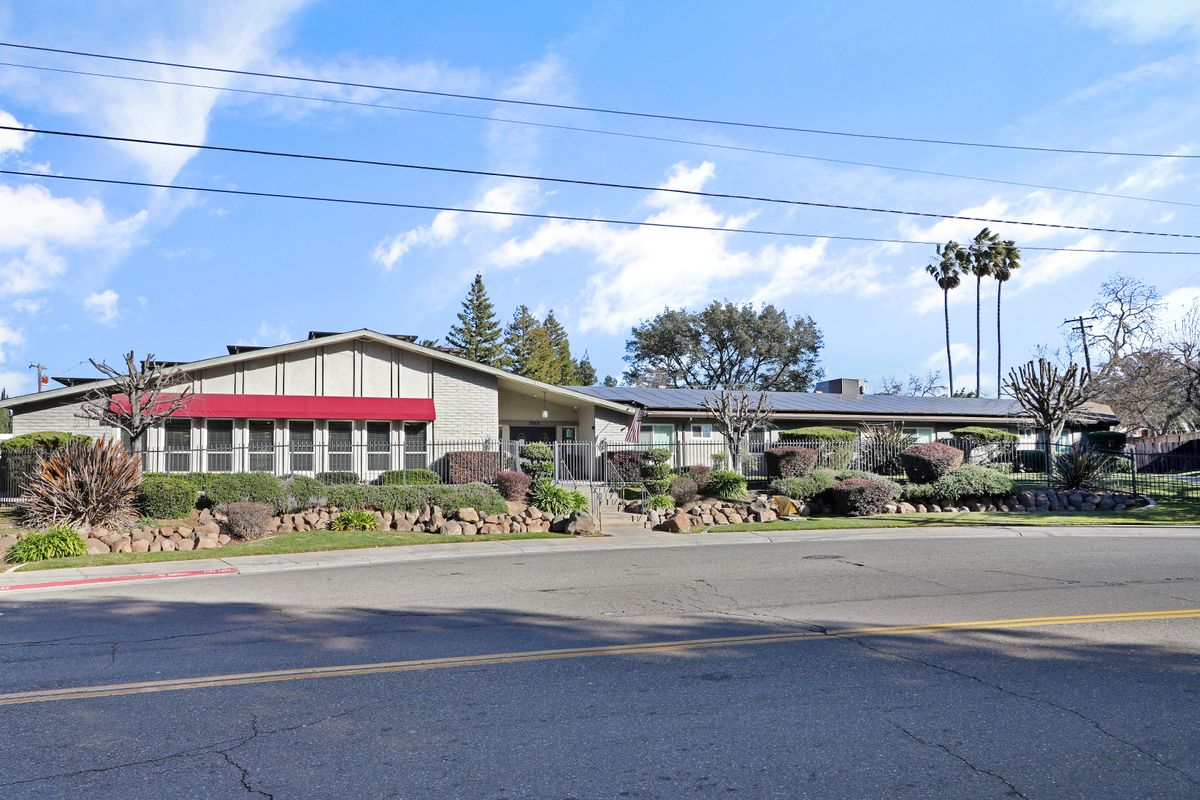 A building with a red awning near a road, surrounded by trees under a blue sky with clouds.