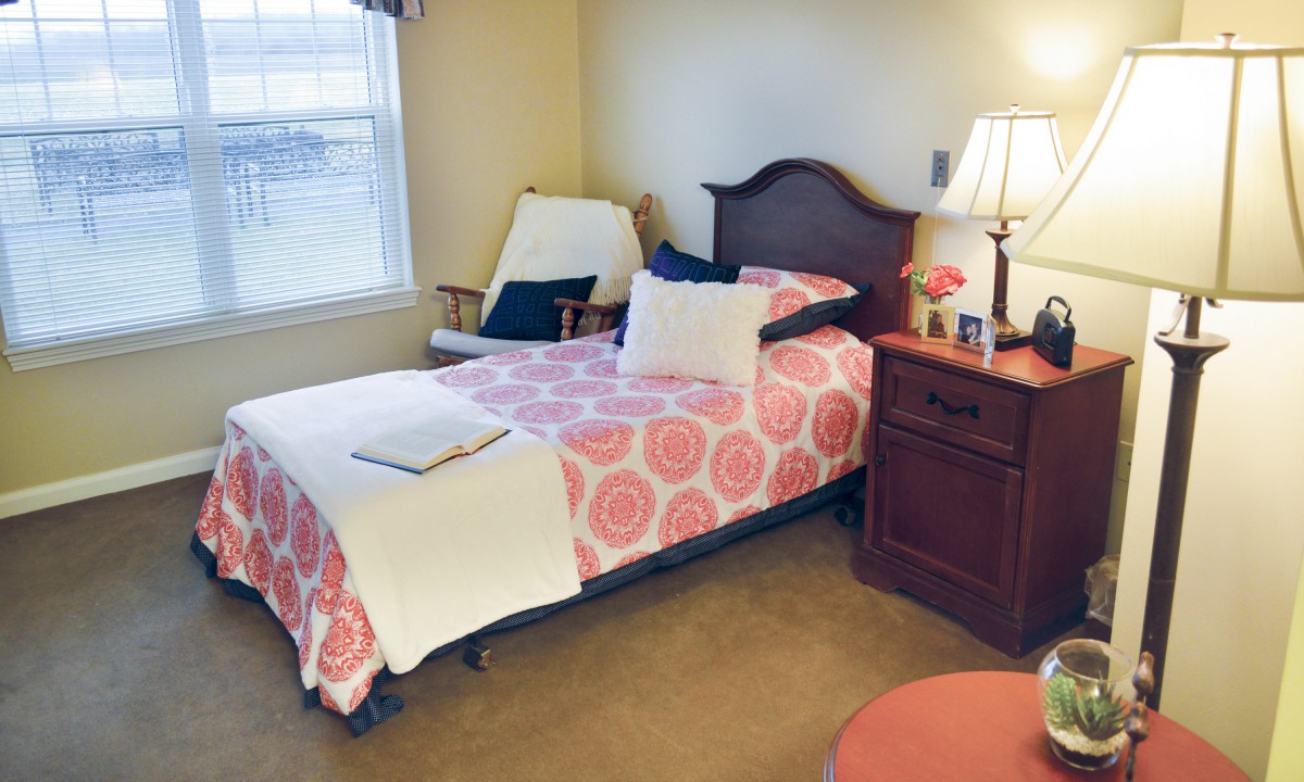 A cozy bedroom with a single bed, featuring a floral bedspread, a wooden nightstand with a lamp, and an armchair by the window.