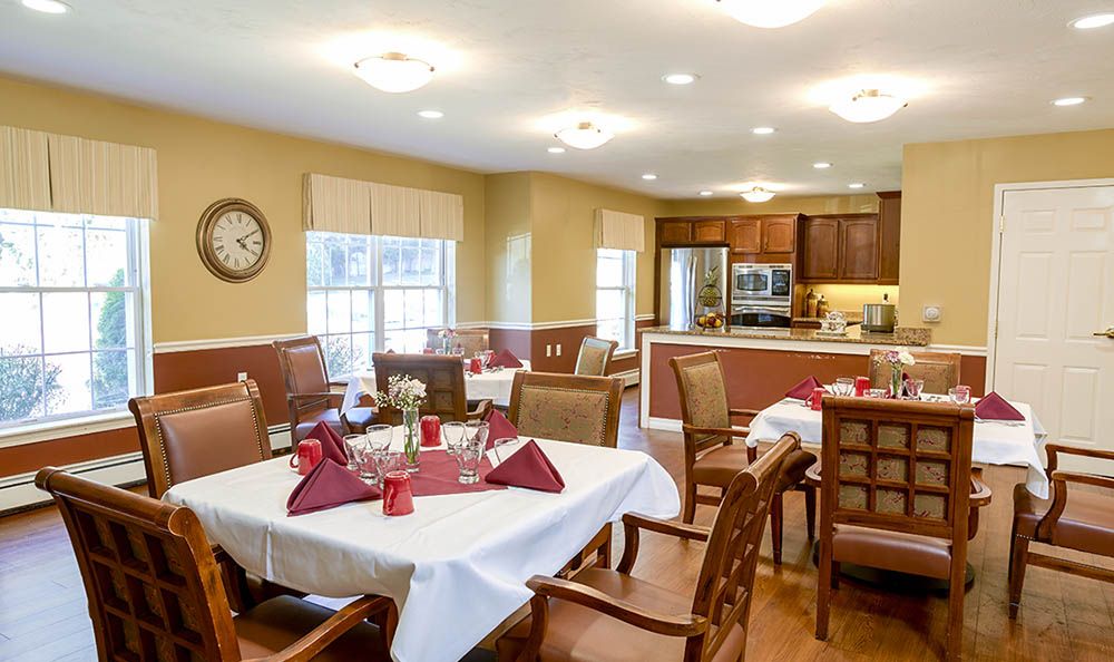 A well-lit dining room with neatly arranged tables and chairs, adjacent to a kitchen area.