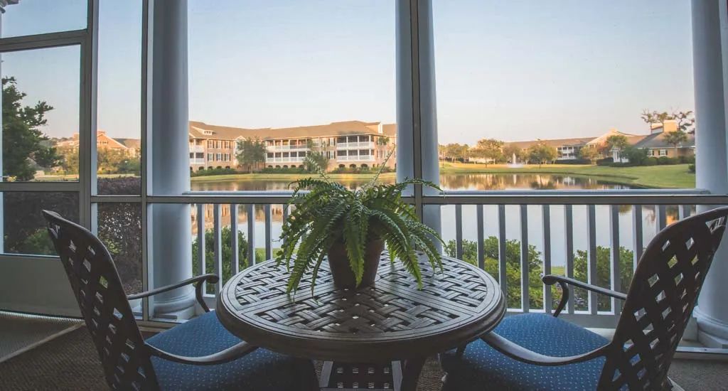 A peaceful balcony scene with a table and plant overlooking a lake and residential buildings.