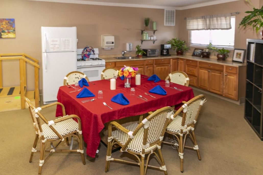 A dining area with a red tablecloth and blue napkins, set for a meal in an adjacent kitchen.