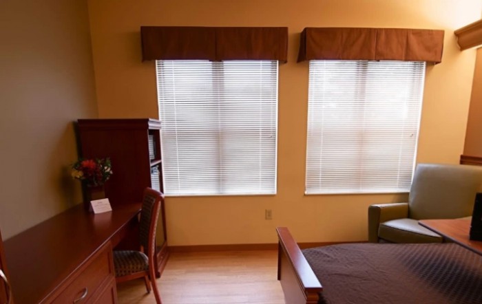 A cozy bedroom with a desk, chair, and large windows with curtains.