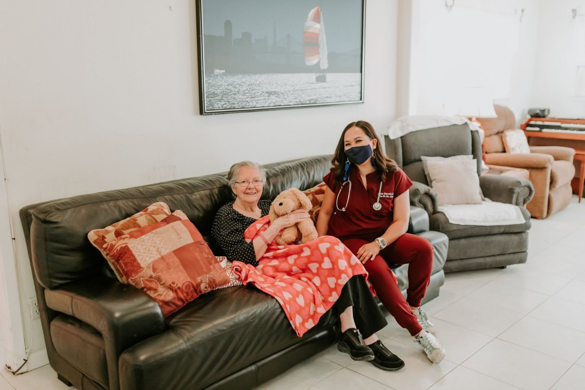 An elderly woman holding a baby sits on a couch with a young woman in a nurse uniform, accompanied by a small dog.