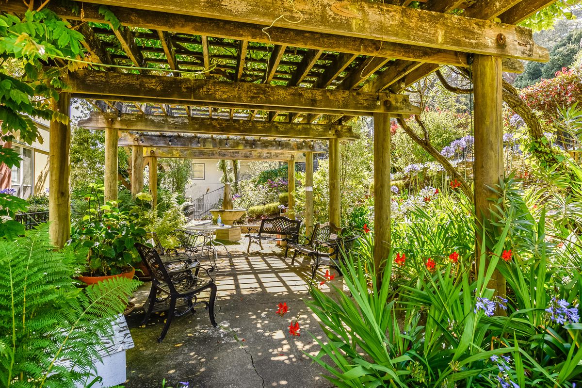 A peaceful garden with a wooden pergola, surrounded by lush greenery and vibrant flowers, bathed in sunlight.