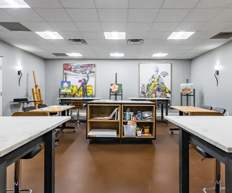 An art studio with easels and paintings displayed, featuring tables and art supplies organized in the center.