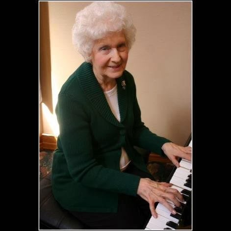An elderly woman playing the piano, dressed in a green jacket and smiling warmly.