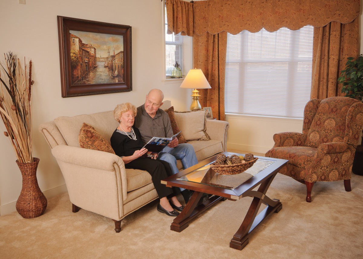 An elderly couple sits together on a sofa in a well-decorated living room, enjoying a book.