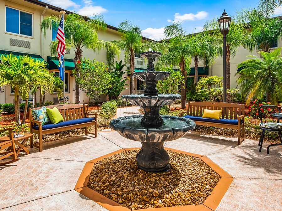 A tranquil courtyard featuring a central fountain surrounded by benches and palm trees.