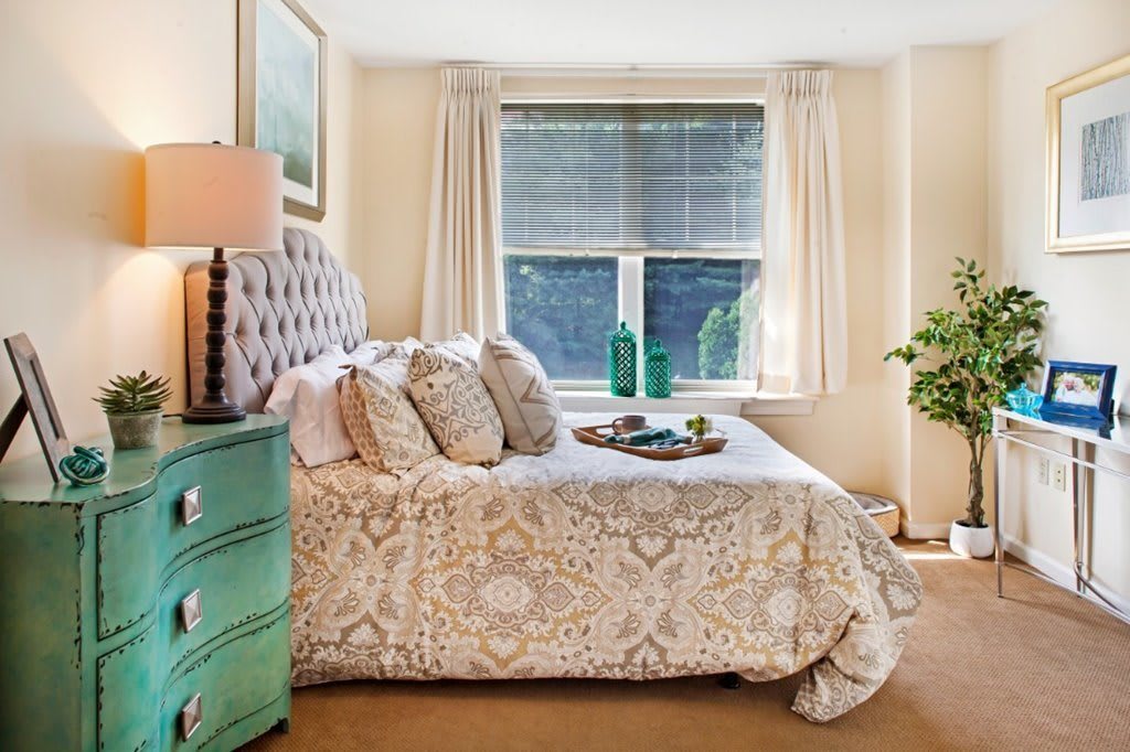 A cozy bedroom with a beautifully decorated bed, featuring patterned bedding and pillows, alongside a green dresser. Natural light flows in through the large window adorned with curtains, complemented by greenery for a fresh touch.