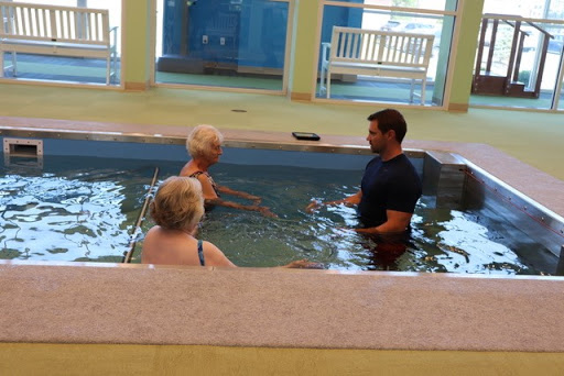 A water therapy session taking place in a swimming pool with a trainer assisting two adults.