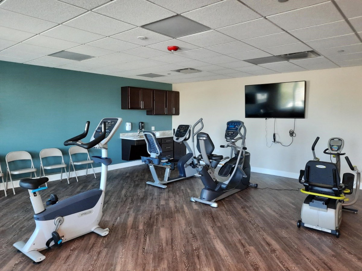 A modern gym with various cardio exercise machines and a large wall-mounted TV.
