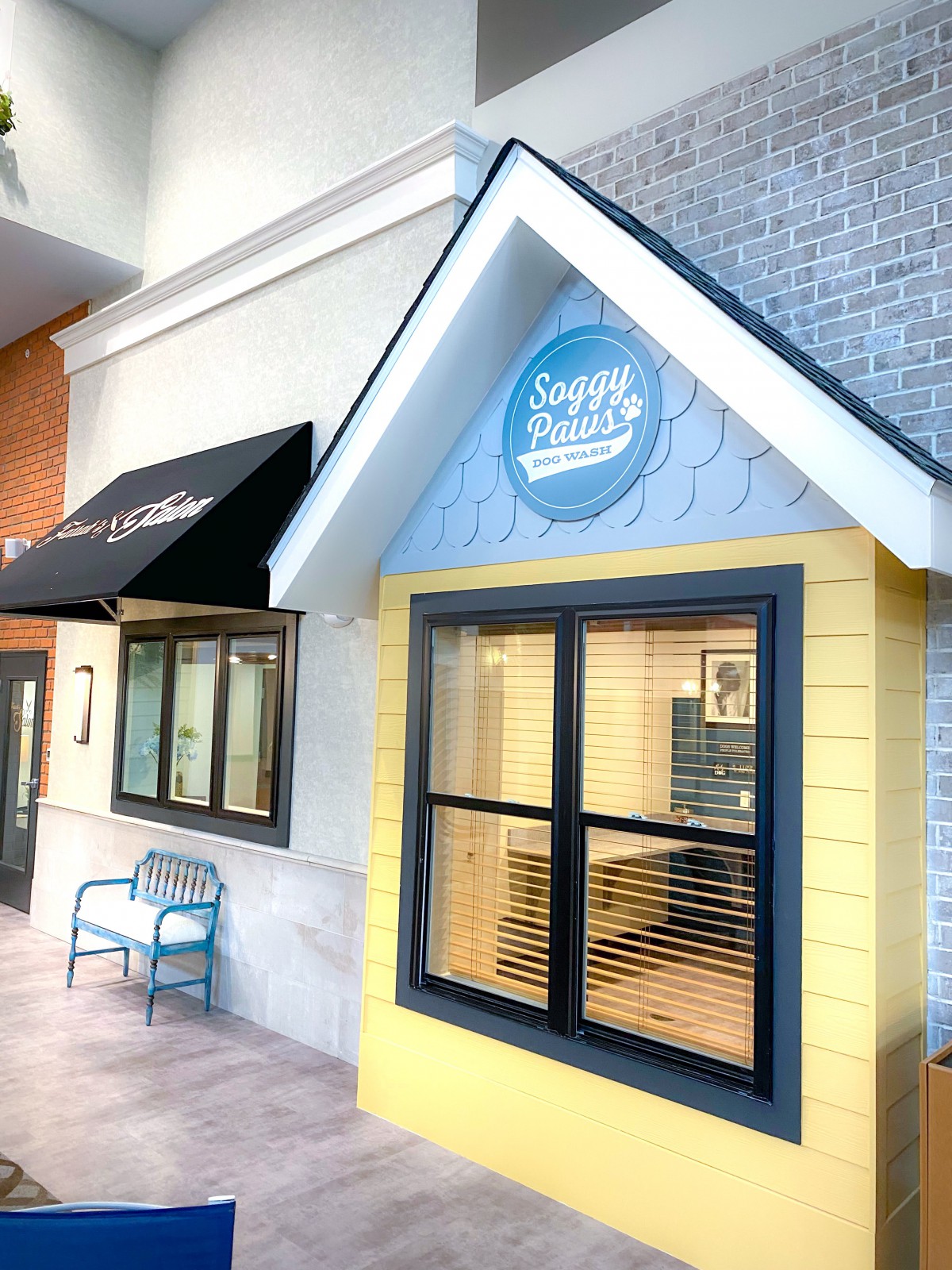 A small storefront with a yellow wall and blue accents, featuring a window, bench, and awning.