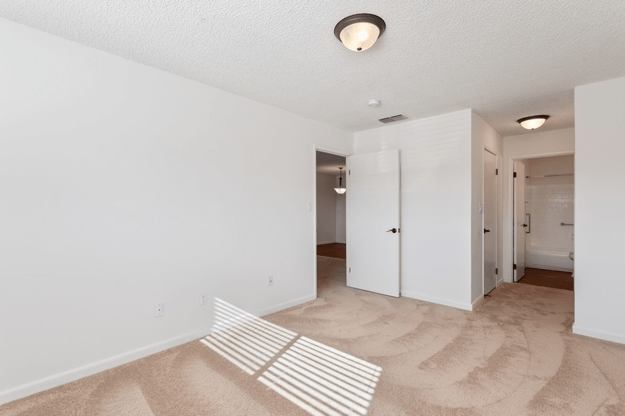 An empty carpeted room with white walls and a ceiling light fixture, featuring an open doorway.