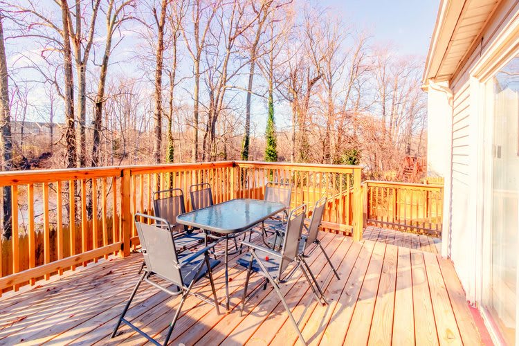 A sunny wooden deck with a glass-top table and chairs, surrounded by trees.